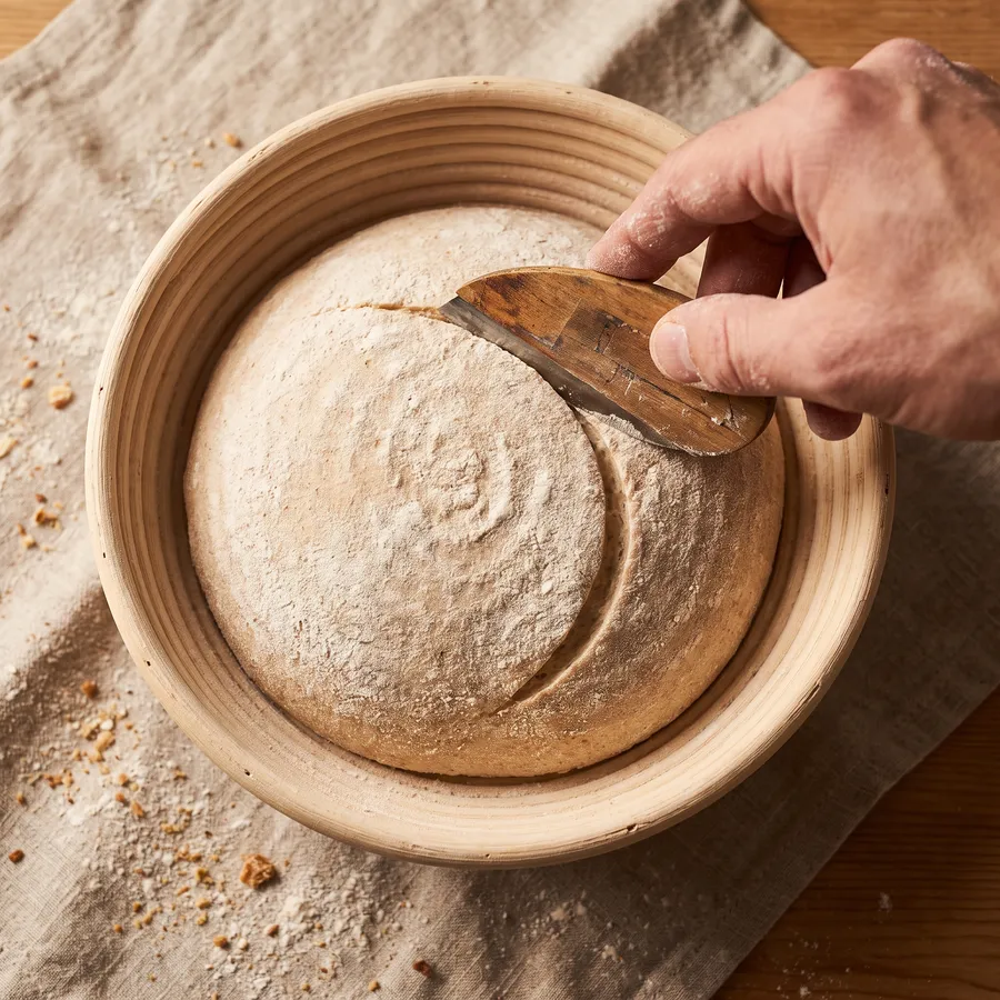 A hand scoring a spiral-floured round of dough in its proving basket
